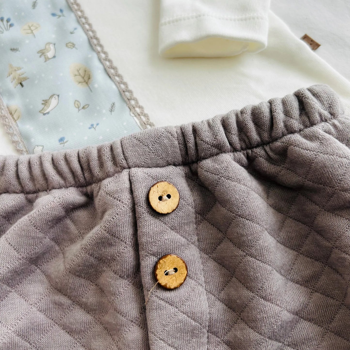 Pormenor de conjunto de menino de camisola branca com faixa decorativa e tapa-fraldas cinza