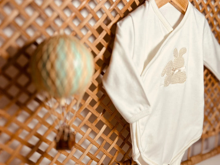 Body de trespasse com collant cardado com motivo coelhos tricot com lacinho na cor pérola pendurado num suporte de vime com um balão de ar quente de brincar ao lado