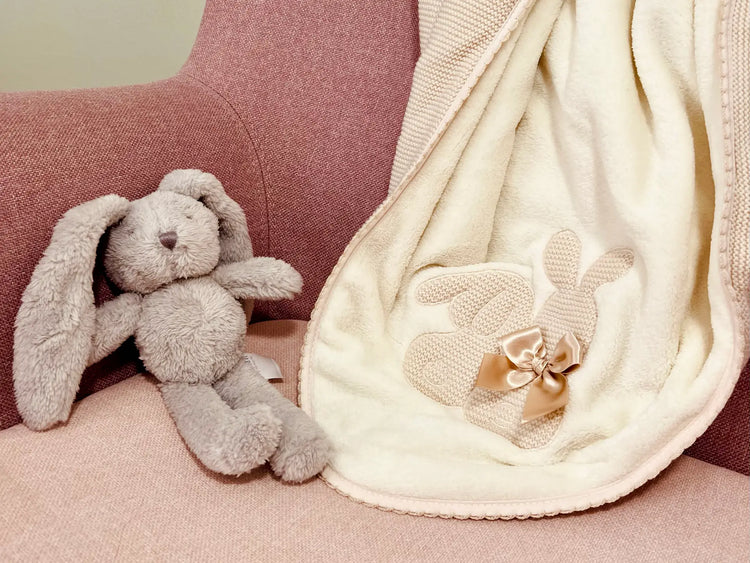 Manta com motivo coelhos tricot com laço na cor bege disposta sobre um cadeirão com um coelhinho de peluche ao lado