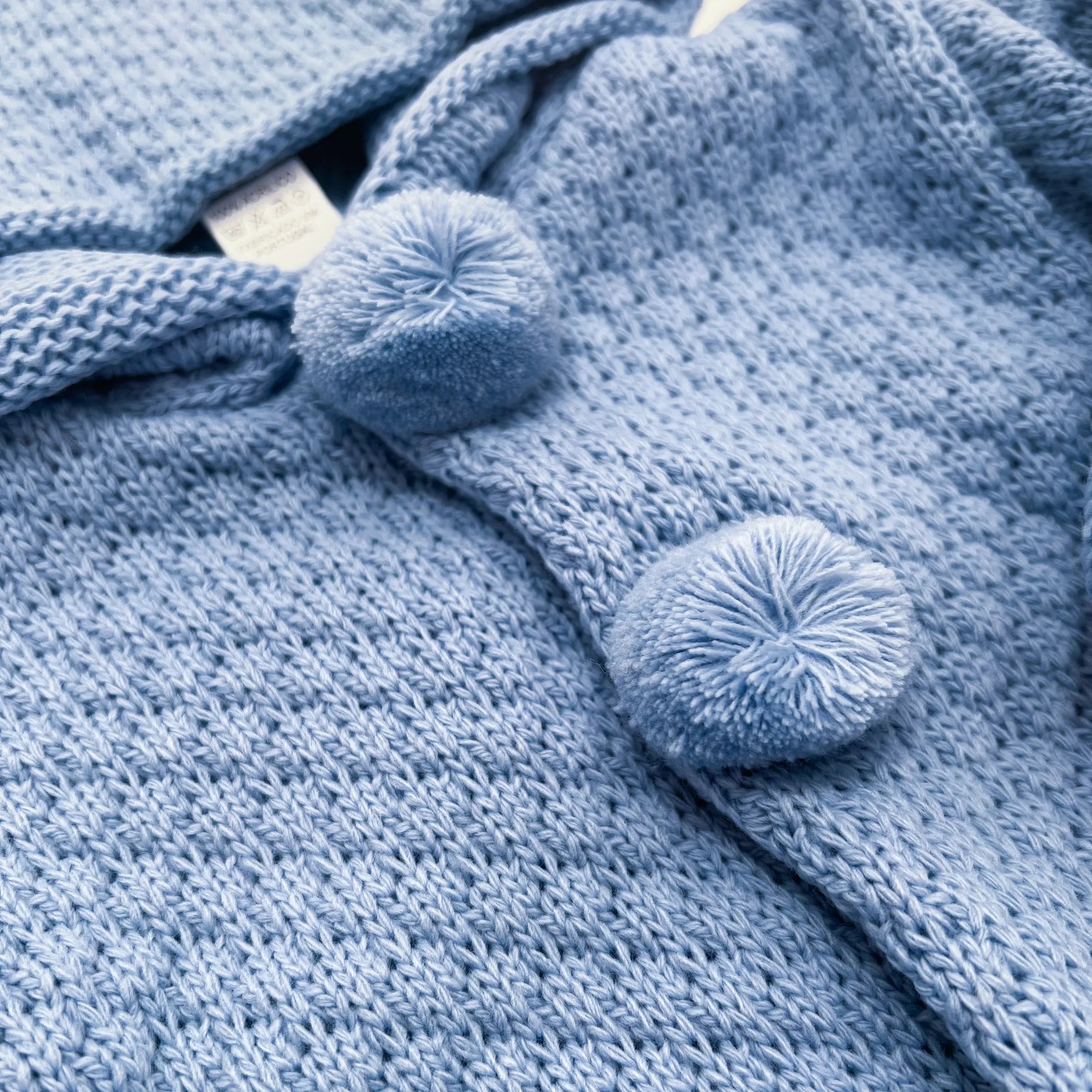 Pormenor de casaco de malha com capuz com pompons na zona dos botões azul