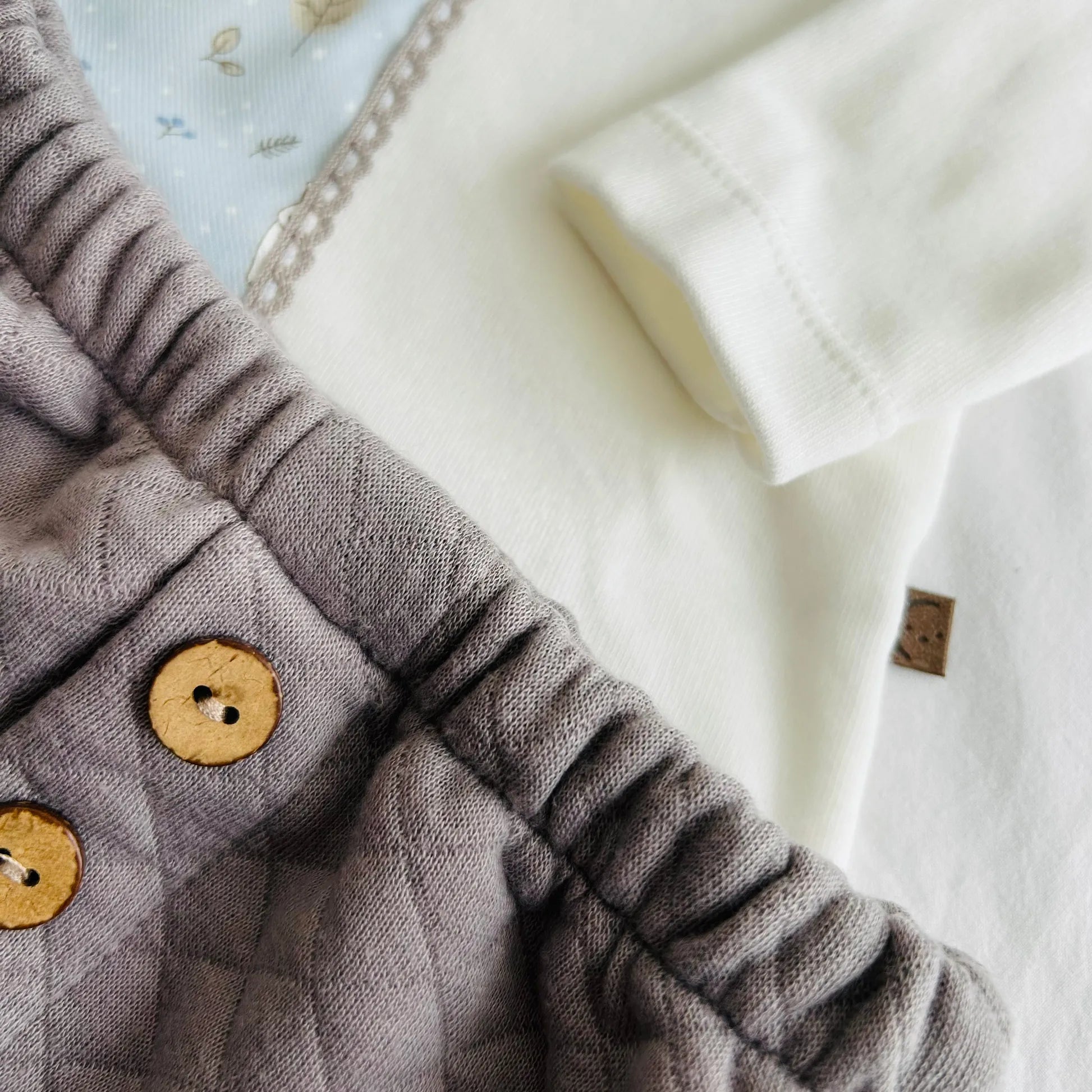 Pormenor de conjunto de menino de camisola branca com faixa decorativa e tapa-fraldas cinza