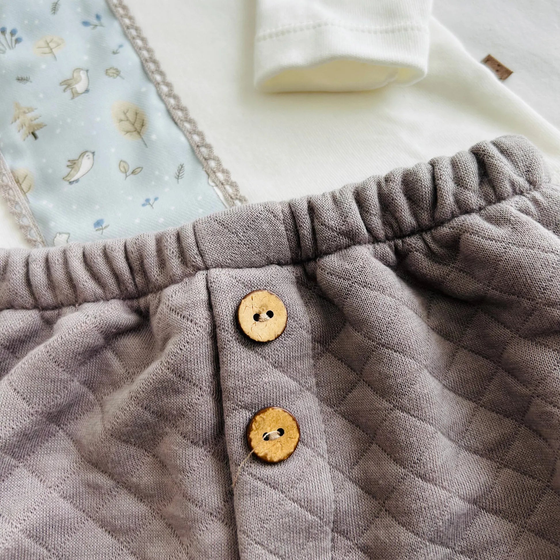 Pormenor de conjunto de menino de camisola branca com faixa decorativa e tapa-fraldas cinza